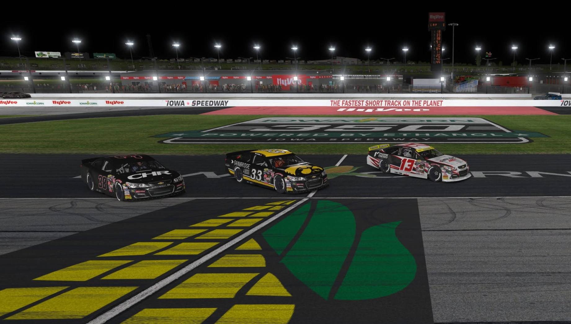 Three oval cars racing at Iowa Speedway under the lights